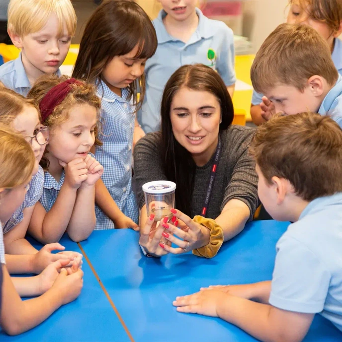 Adult seated at a classroom table surrounded by a group of pupils, holding a clear container as part of a hands-on learning activity. Pupils gather closely around the table observing the demonstration, with classroom furniture and educational resources visible in the background, highlighting an interactive and collaborative learning environment.