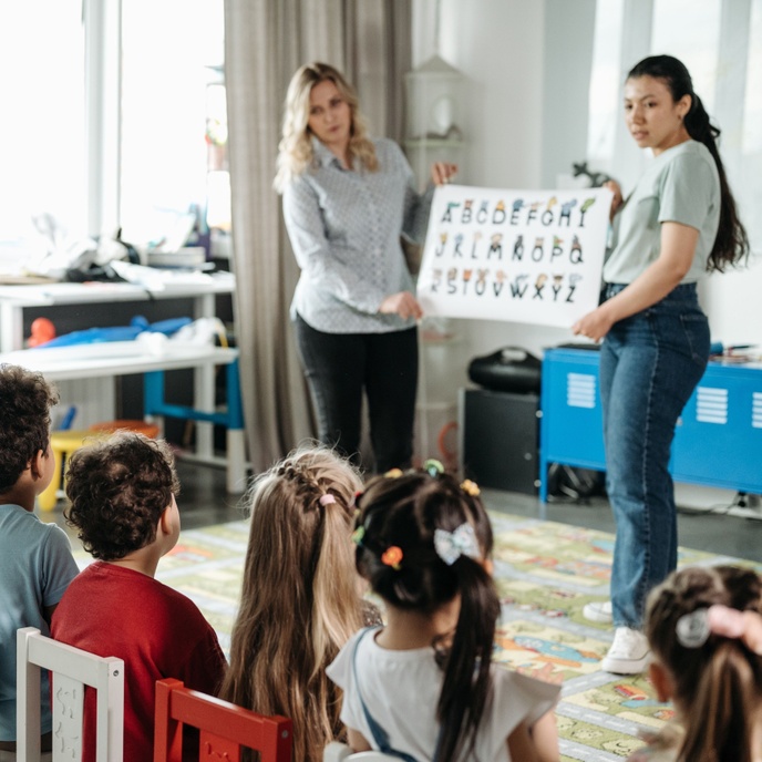 Adults presenting an alphabet chart to a group of seated pupils during a classroom learning activity, supporting early literacy and letter recognition skills.