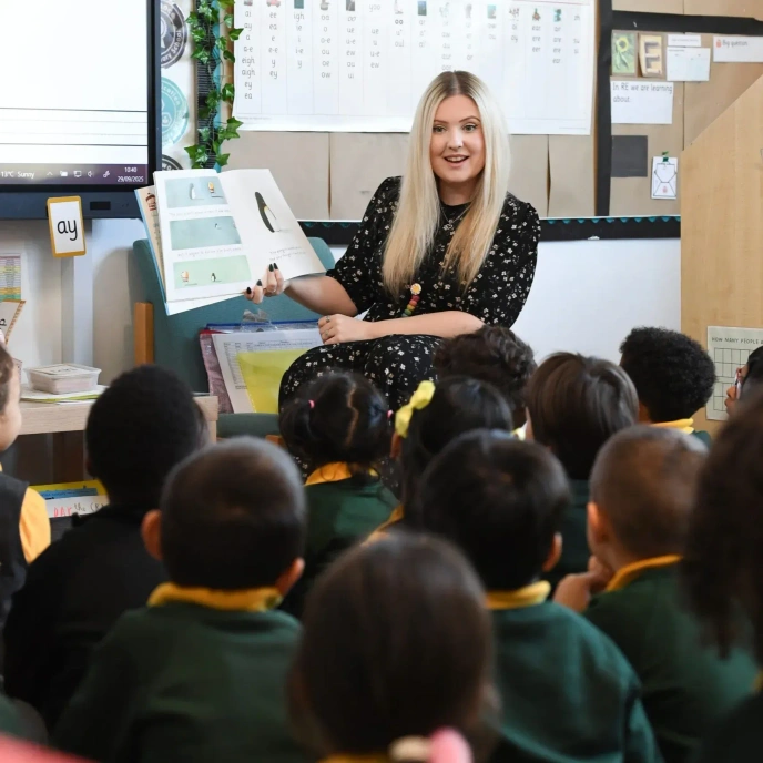 An adult reading a book to a group of primary school pupils seated on cushions in a classroom, with bookshelves and learning resources in the background.