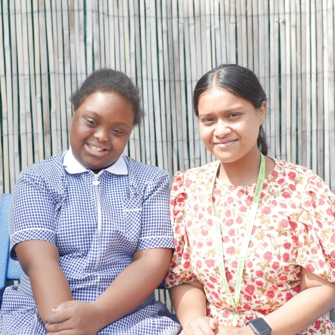 A pupil and an adult seated outdoors side by side, looking towards the camera in front of a fence backdrop.