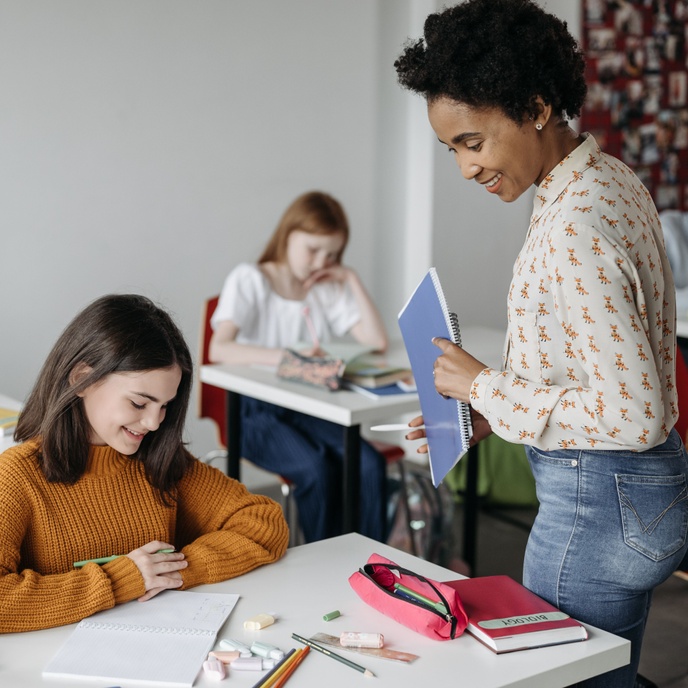 Adult supporting a pupil during classroom learning, providing guidance at a desk while other pupils work independently in the background.