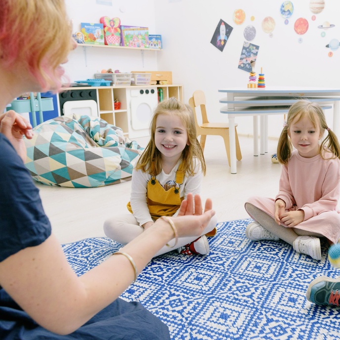 Adult leading a group learning activity with pupils seated on a classroom rug, supporting communication, listening and early years development skills.