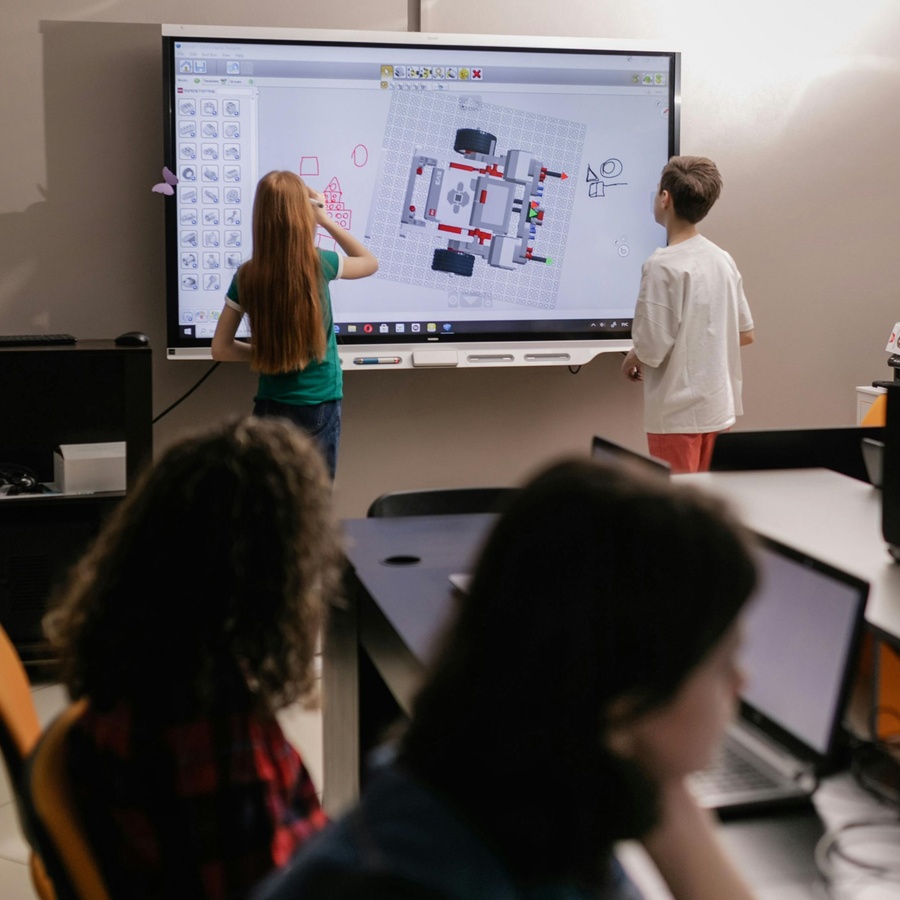 Children standing at an interactive whiteboard in a classroom, drawing or arranging shapes on a large digital display, while other students sit at desks using computers in the foreground.