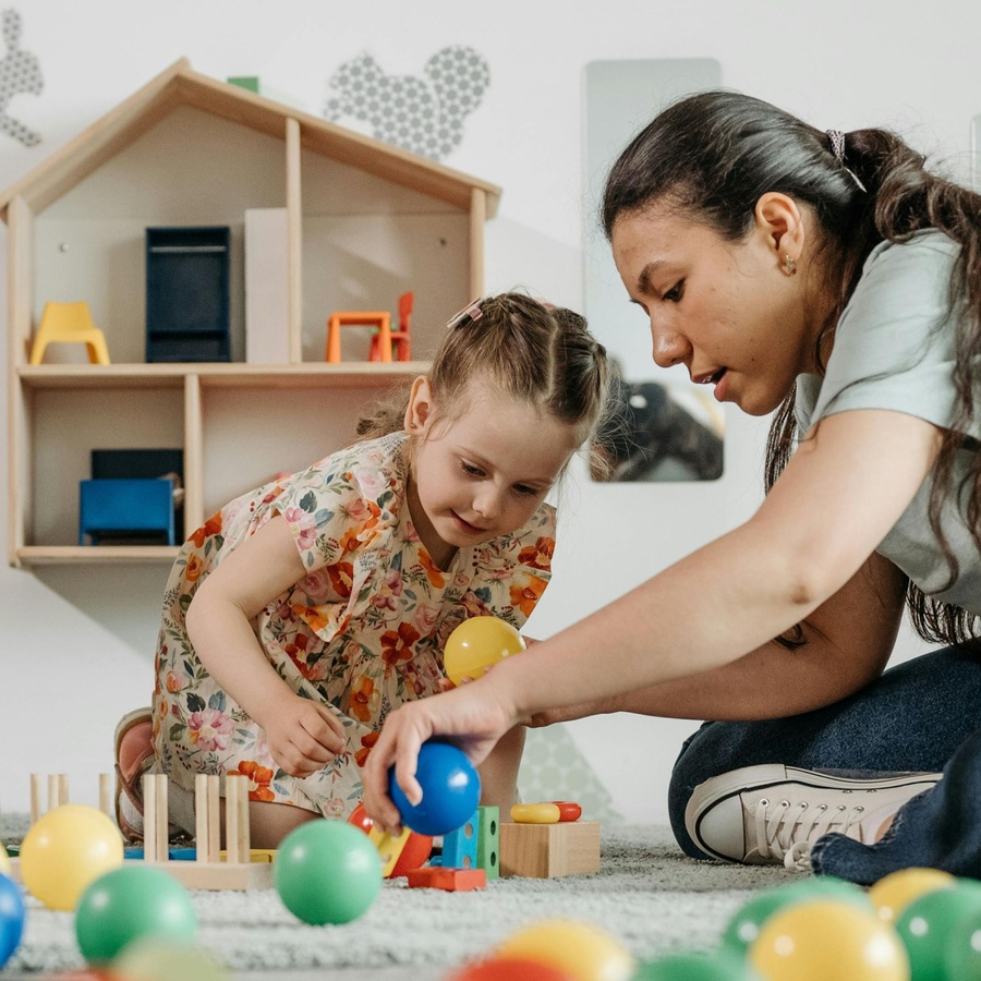 Adult and young child sitting on the floor indoors, playing together with colourful wooden toys and balls in a childcare or playroom setting.