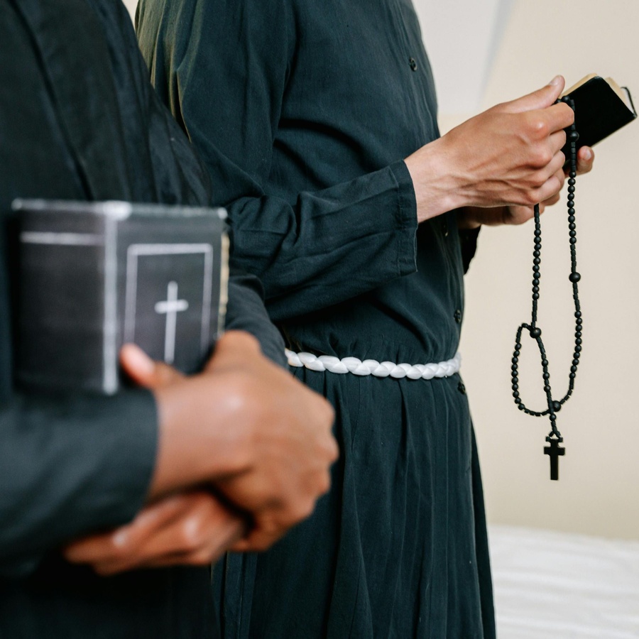 Two people wearing long dark robes standing indoors, one holding a book with a cross symbol on the cover and the other holding a rosary with a cross.