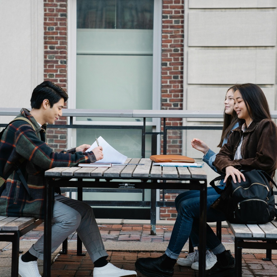 Two people seated at an outdoor table, engaged in conversation, with papers and a notebook on the table and a brick building in the background.