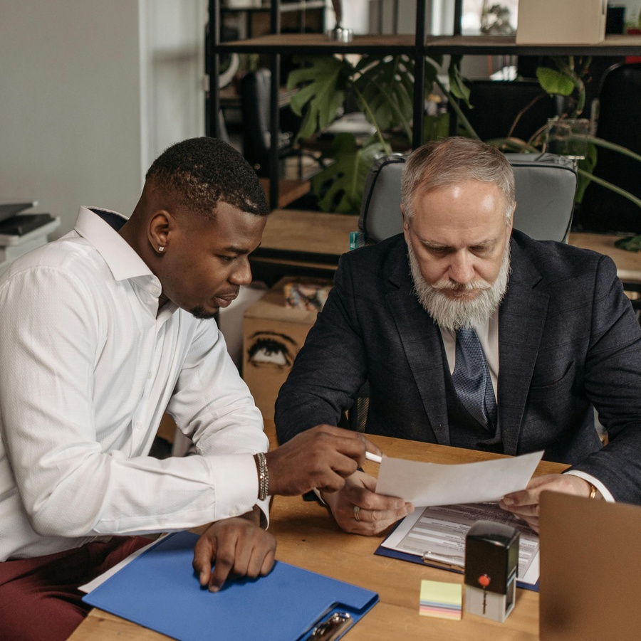 Two people seated at a desk in an office setting, reviewing documents together with folders and papers spread out on the table.