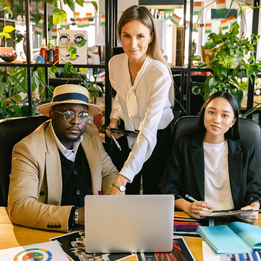 Three people seated and standing around a desk in a modern office, reviewing information on a laptop with notebooks, documents, and colourful charts spread out on the table, surrounded by indoor plants and shelving.