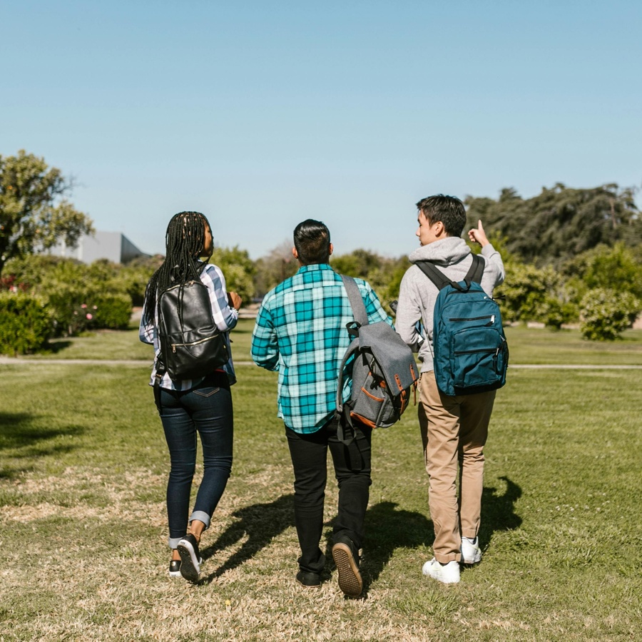 Three people walking across a grassy outdoor area with backpacks, heading away from the camera toward buildings and trees.