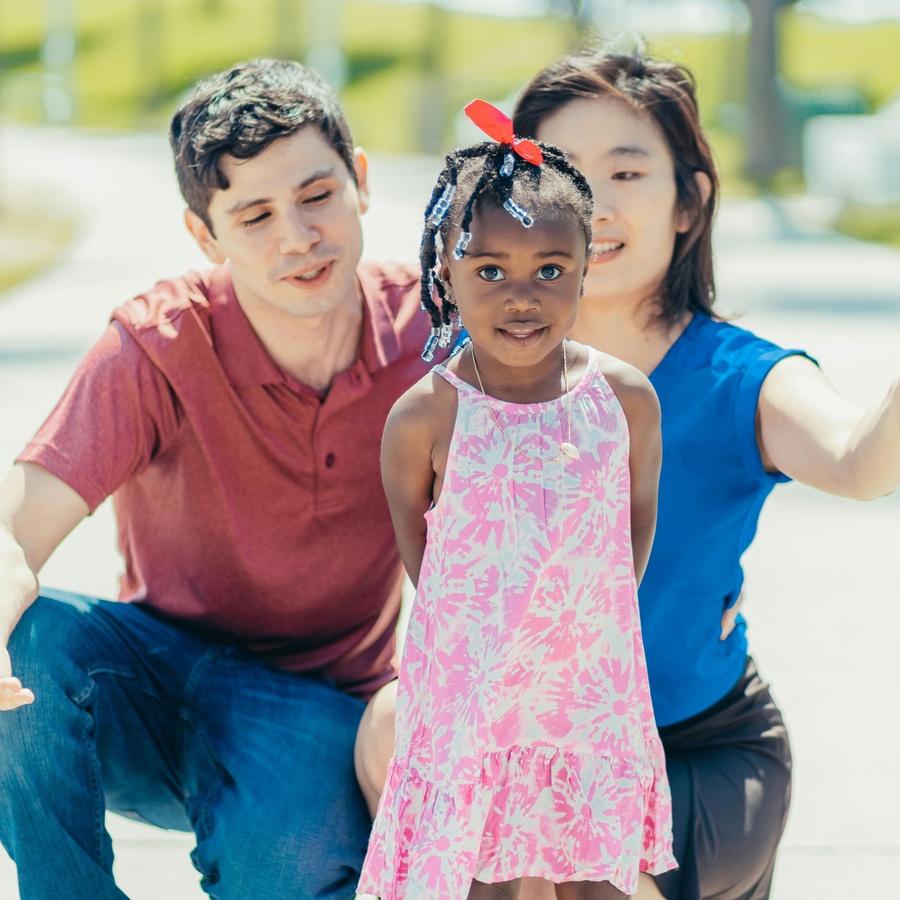 Young child standing outdoors in a pink patterned dress, with two adults crouched slightly behind and reaching forward in a bright, sunny setting.