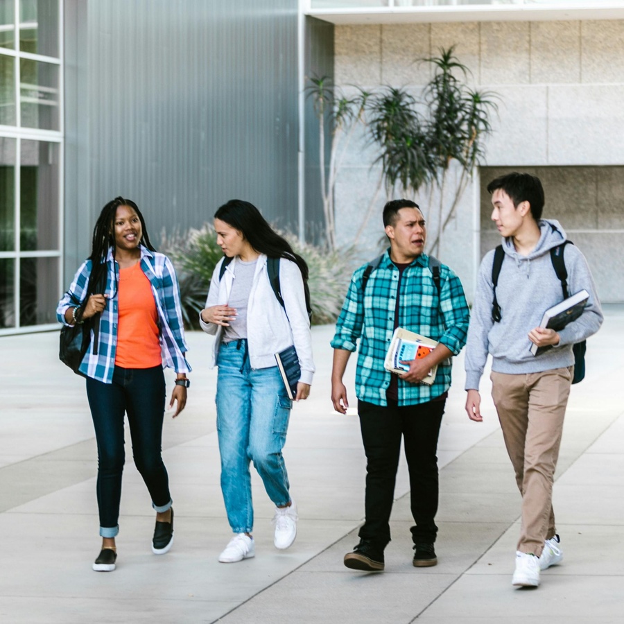 Four people walking together outdoors on a campus pathway, carrying backpacks and books, with modern buildings in the background.