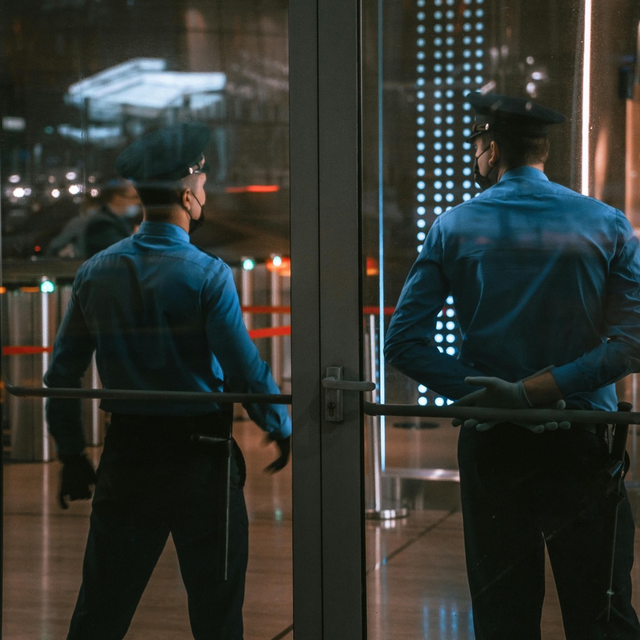 Two uniformed personnel standing inside a building near glass entrance doors, viewed from behind, with reflective surfaces, city lights, and illuminated panels visible through the glass in a modern indoor setting.