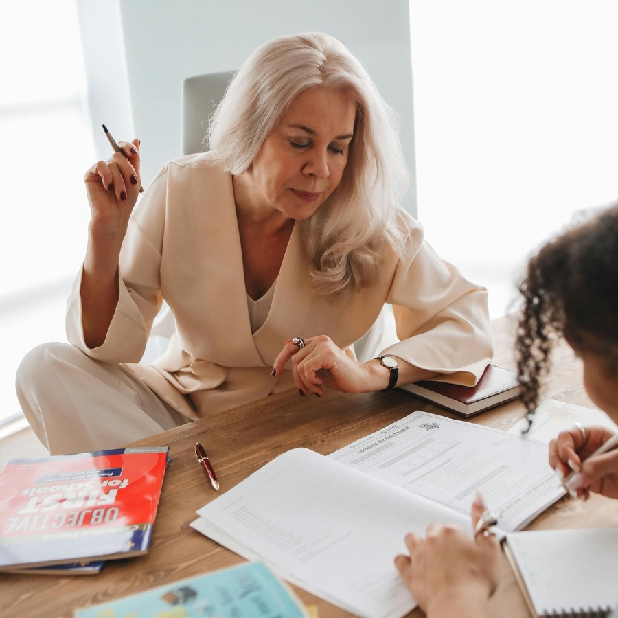 Two people sit at a table reviewing open books and documents, with one person holding a pen during a study or work session.