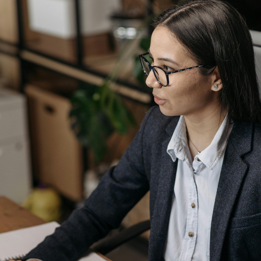 An individual seated at a desk in an office workspace, wearing a blazer and collared shirt, with shelves and plants visible in the background.