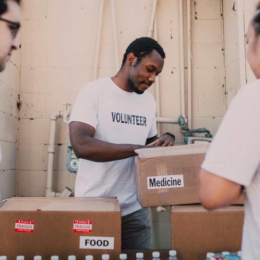 People packing and labelling cardboard boxes of food and medicine in an indoor space, with one person wearing a “Volunteer” T‑shirt handling a box marked “Medicine.”