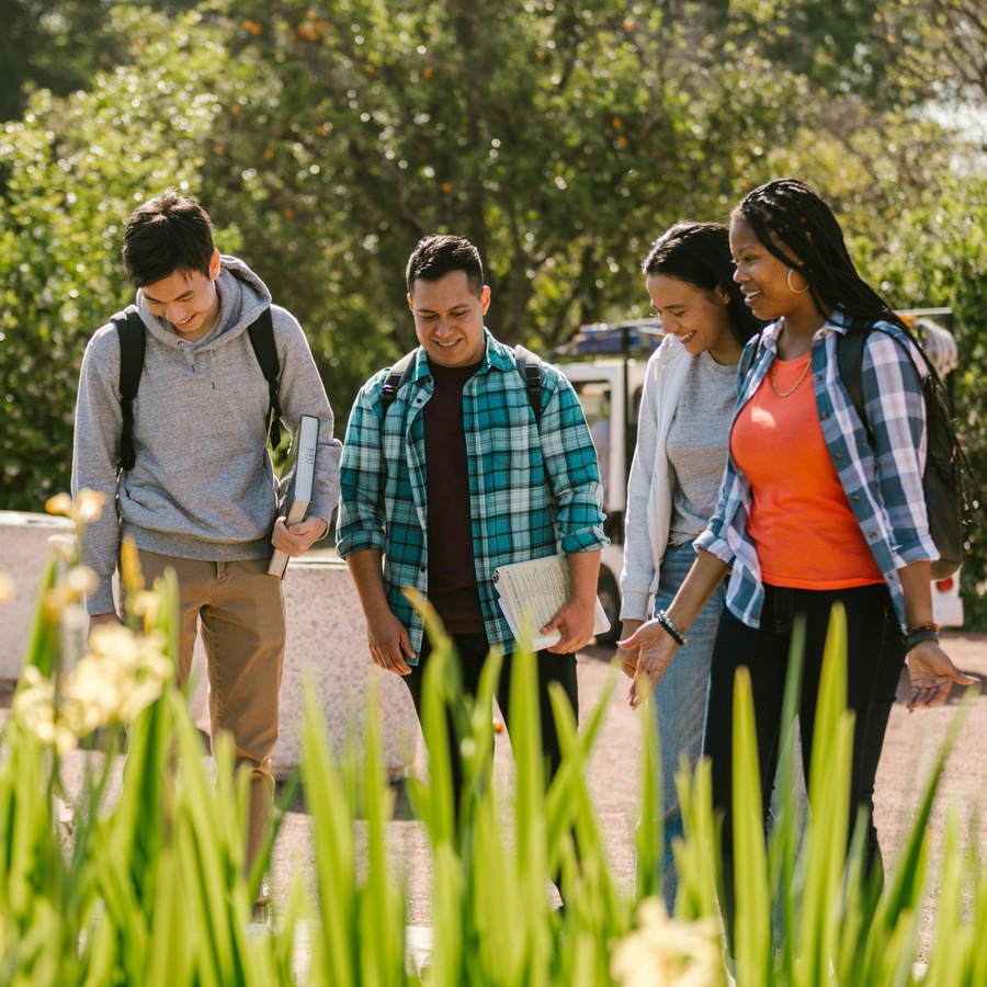 Group of people walking outdoors on a pathway, carrying backpacks and notebooks, with greenery and flowers in the foreground.