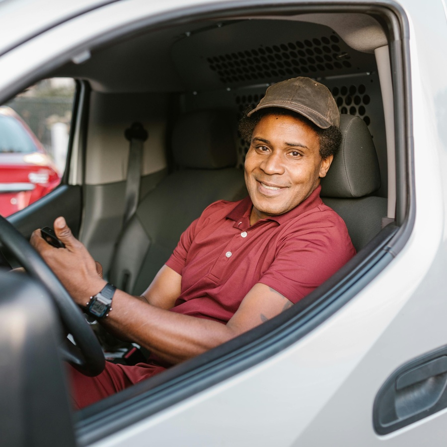 Person seated in the driver’s seat of a vehicle, smiling toward the camera while holding the steering wheel, with the interior of the van and parked cars visible outside.