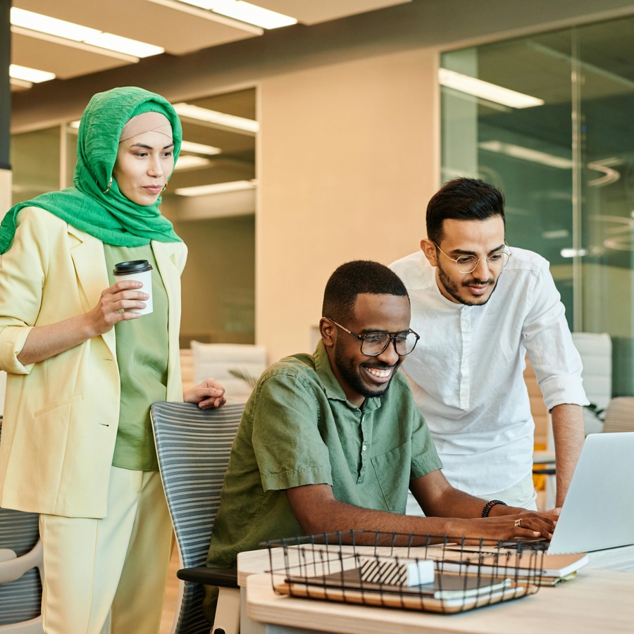 Three people standing around a desk in a modern office, looking at a laptop together, with a takeaway coffee cup, folders, and office shelving visible nearby.