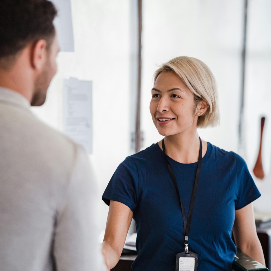 Person standing in an office setting, extending a hand in greeting while wearing a lanyard, with another person partially visible in the foreground.