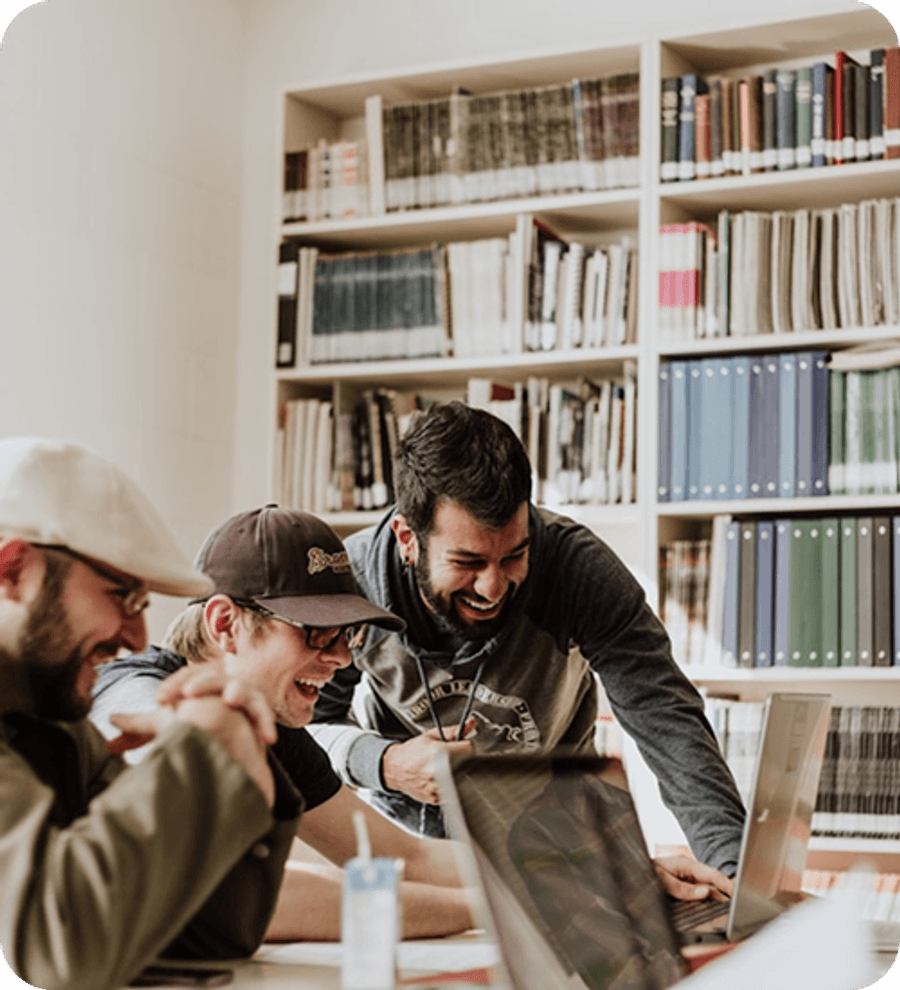 Group of people collaborating at a table in a library, looking at open laptops with bookshelves in the background.