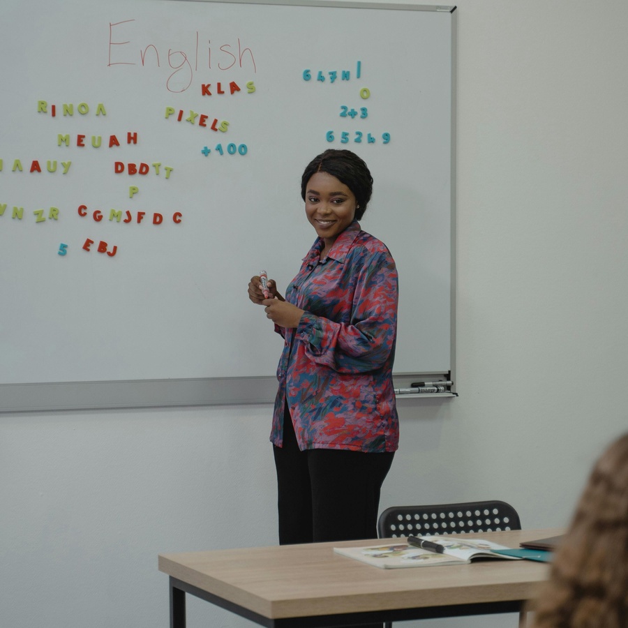 An individual stands at a whiteboard holding a marker, with colorful magnetic letters arranged across the board and a desk with papers in the foreground.