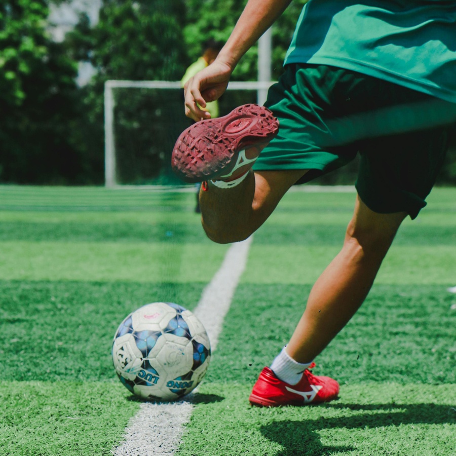 Close‑up of a person kicking a football on a grassy pitch, with one foot raised mid‑strike and the ball positioned near a white sideline on an outdoor sports field.