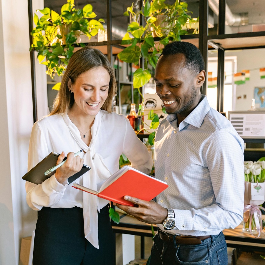 Two people standing in a bright indoor workspace, reviewing a red notebook together while holding a tablet and pen, with indoor plants and shelving visible in the background.