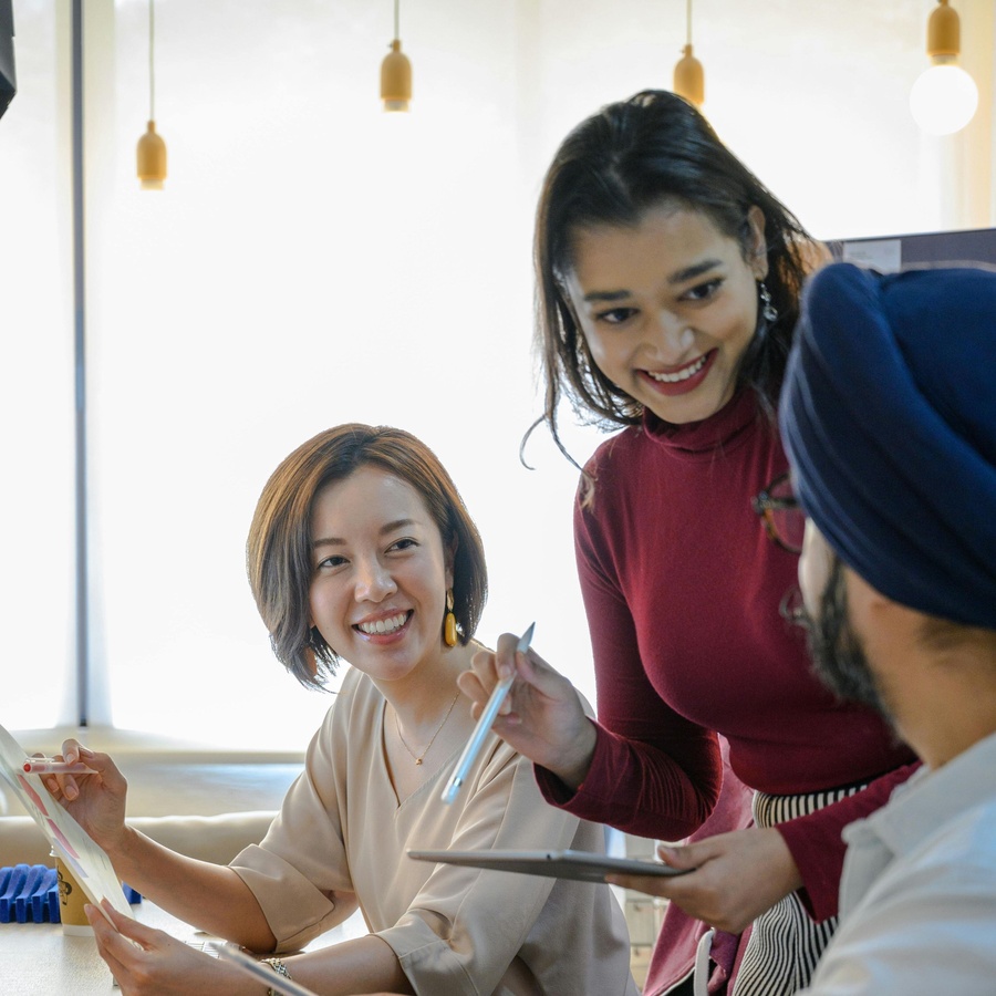 Three people gathered at a table indoors, smiling and pointing at documents while one person holds a clipboard and another uses a pen, with bright natural light in the background. 