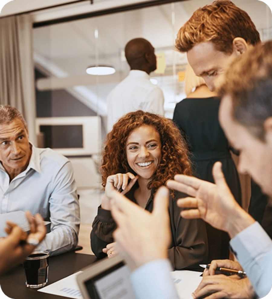 A group of people seated around a table in a busy office setting, engaged in discussion while others work in the background.
