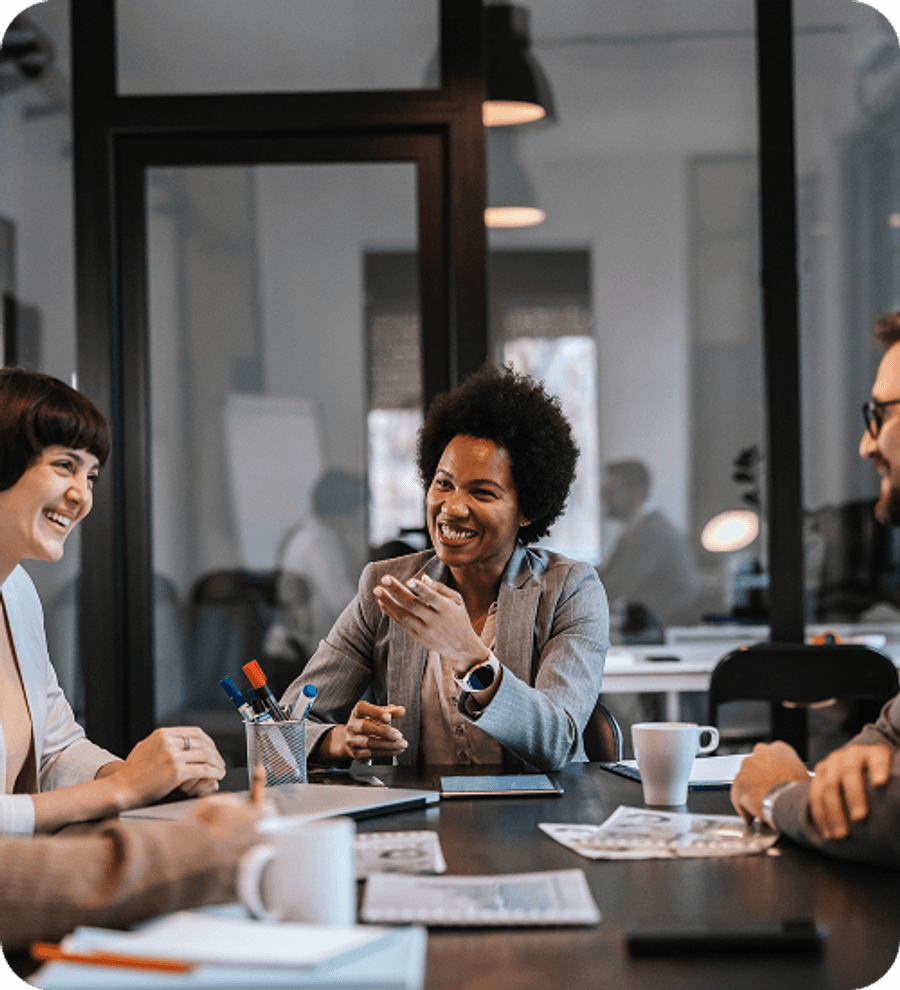 Several people sit around a conference table in a modern office, talking and taking notes while one person gestures during the discussion.