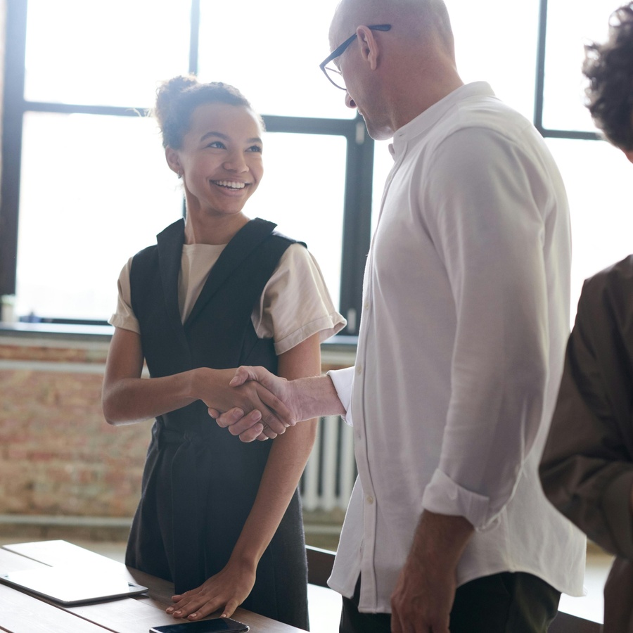 Two people shaking hands across a table in a bright office space, with documents on the table and other people standing nearby in the background.