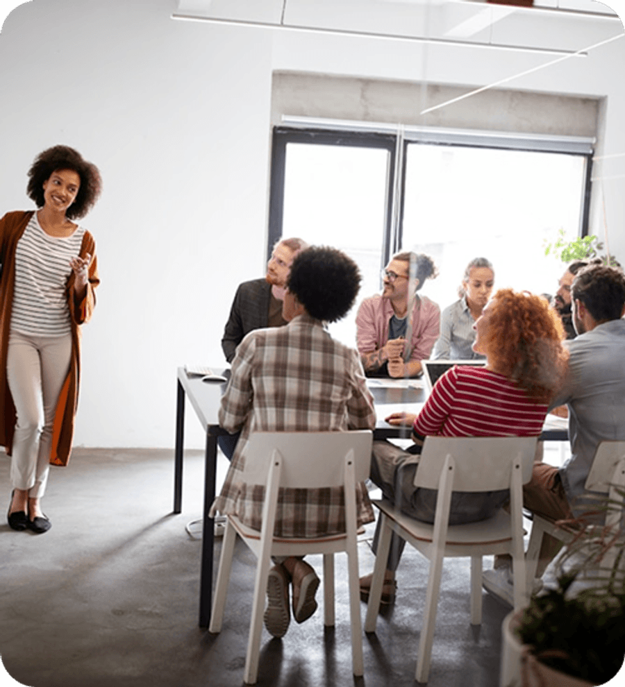A group of people sit around a meeting table listening to a person standing at the front of the room near large windows.