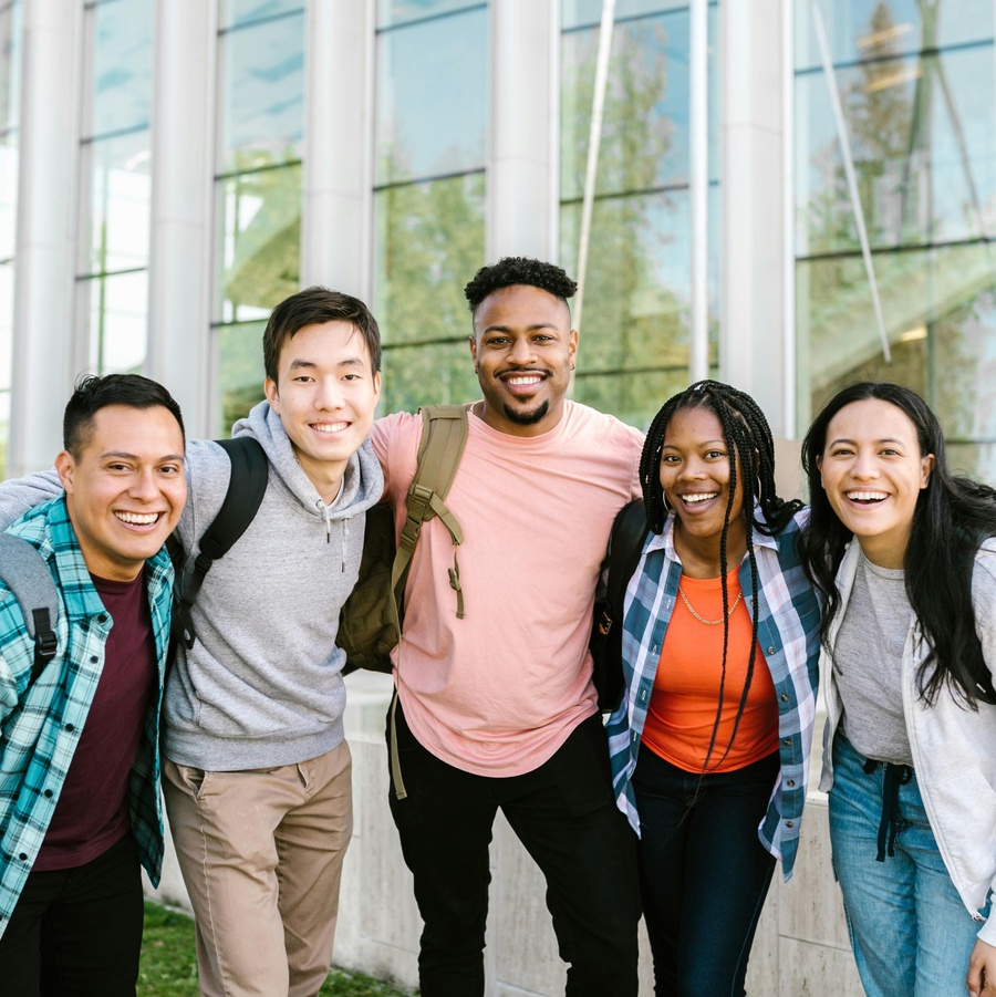 Group of people standing closely together outdoors in front of a glass building, wearing casual clothing and backpacks.