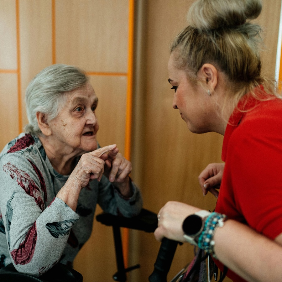 Older adult seated indoors, leaning forward and gesturing with hands while speaking to a standing caregiver who is holding mobility aids, in a warmly lit care or residential setting with wooden walls in the background.