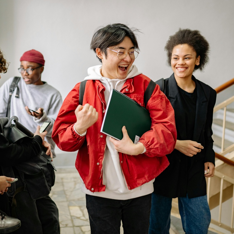 Group of people walking indoors, with one person holding a notebook and wearing a backpack, while others walk nearby carrying bags and phones.