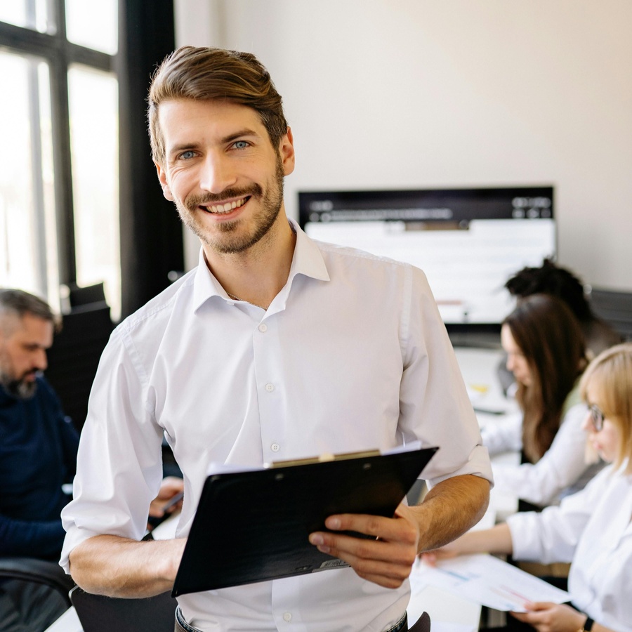 Person standing in a modern office, holding a clipboard and smiling toward the camera, with colleagues seated and working at desks in the background.
