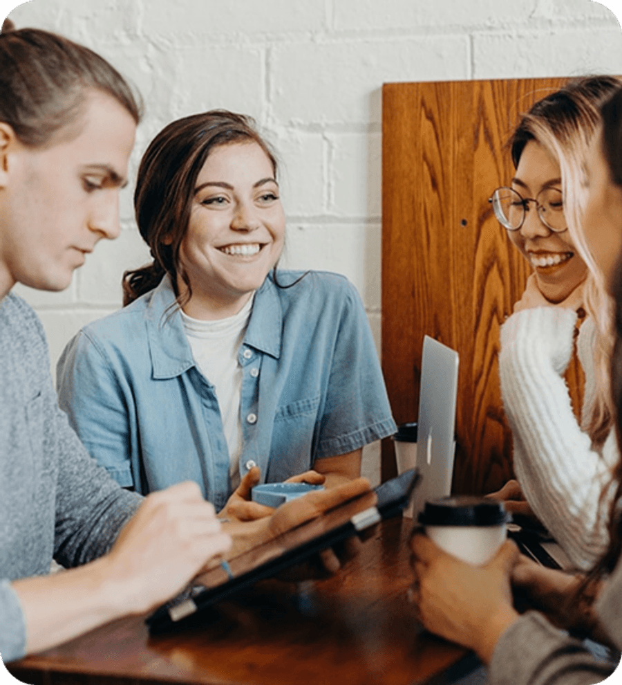 Group of people sitting closely together at a table indoors, using a tablet and mobile phone, with coffee cups and notebooks on the table.