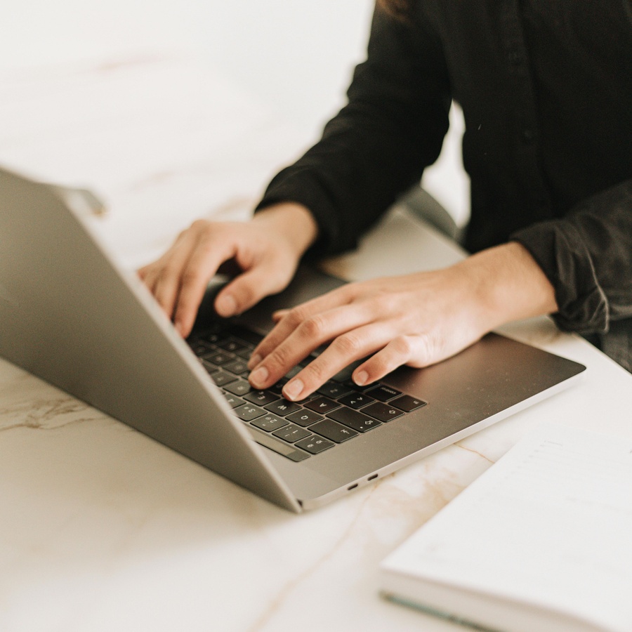 Hands typing on a laptop on a light-colored desk, with a notebook nearby.