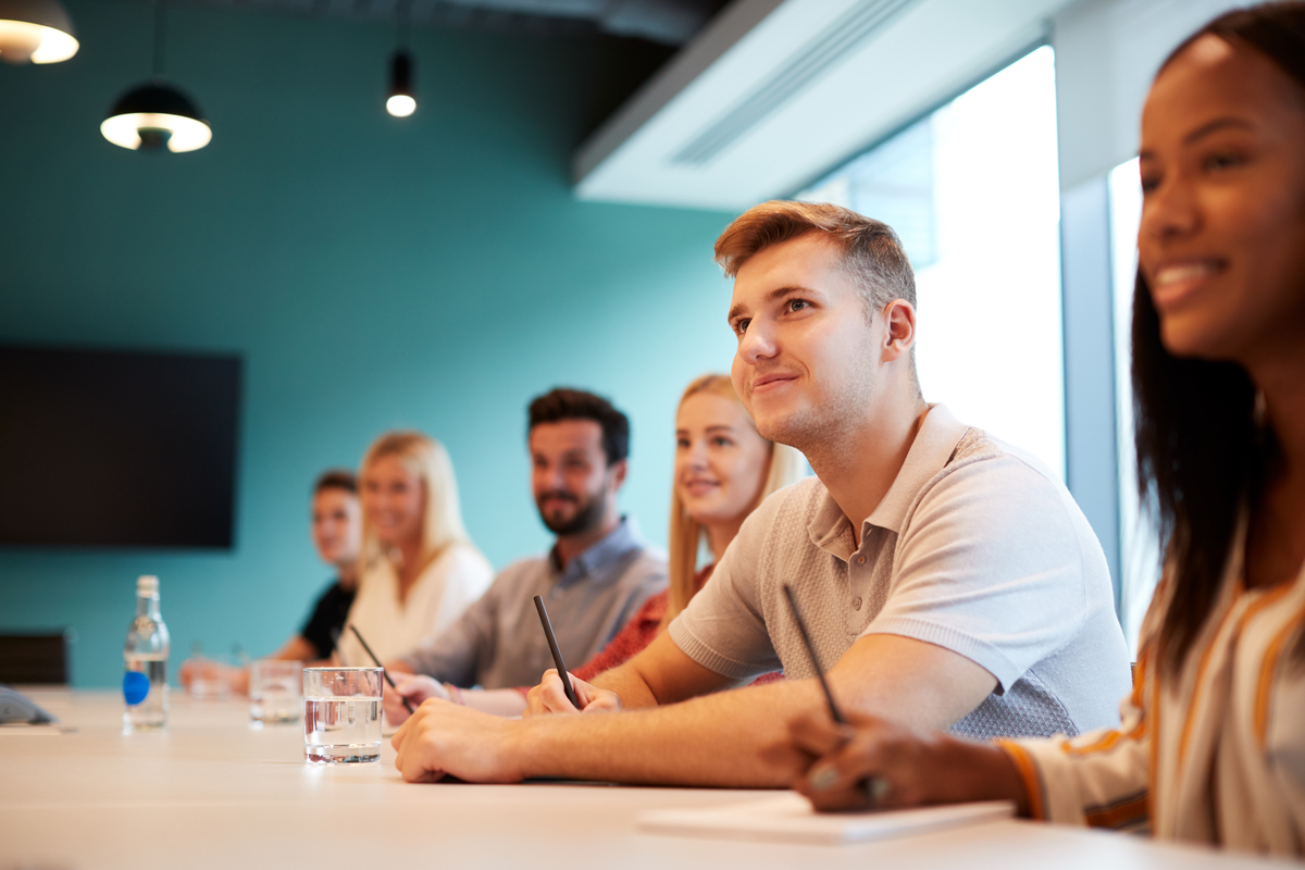  A diverse group of young professionals sits at a long conference table, listening attentively to a presentation. The person in the foreground smiles while taking notes, with a glass of water and a bottle next to them on the table.