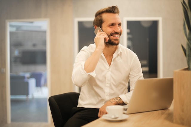  A man with a beard and a white shirt is sitting at a desk and smiling while talking on a cell phone. There is an open laptop and a cup of coffee on the desk in front of him.