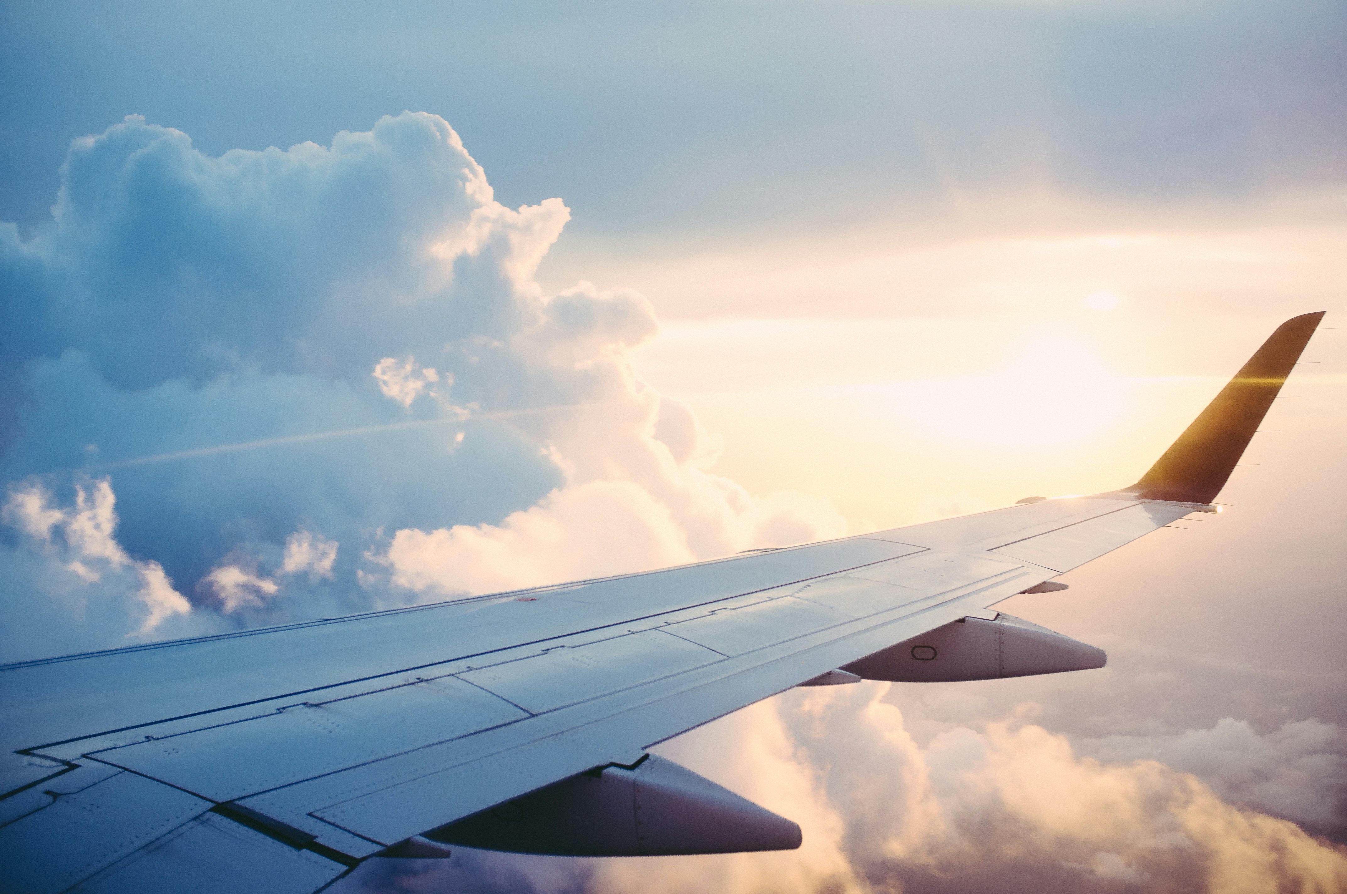 Airplane wing cutting through golden sunrise light above a sea of clouds, viewed from a window seat.