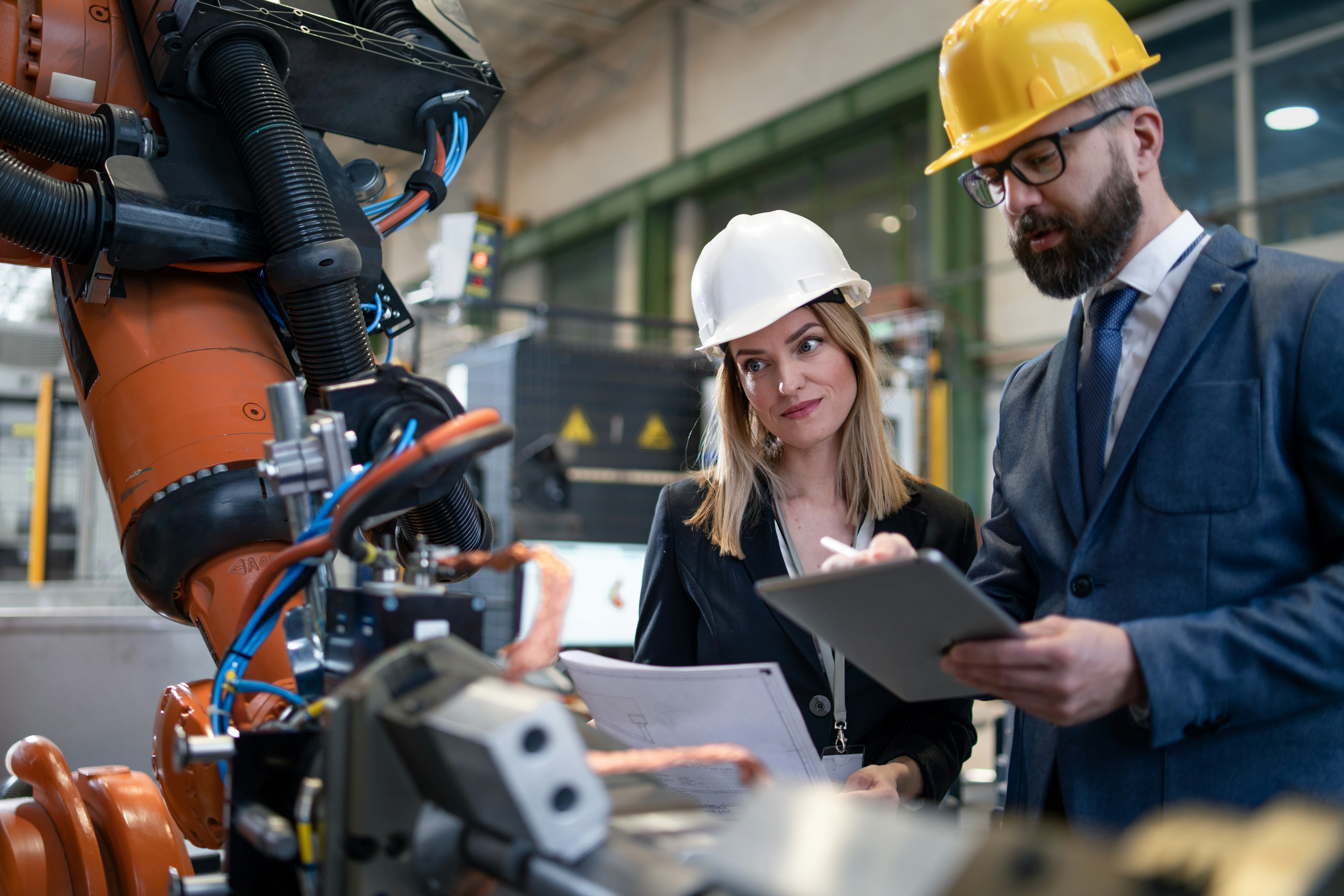 A woman and a man wearing hard hats stand next to an industrial robotic arm in a factory. The woman holds technical documents and looks thoughtfully at the machine, while the man in a suit and yellow helmet reviews notes on a clipboard. Cables and robotic components are visible in the foreground.