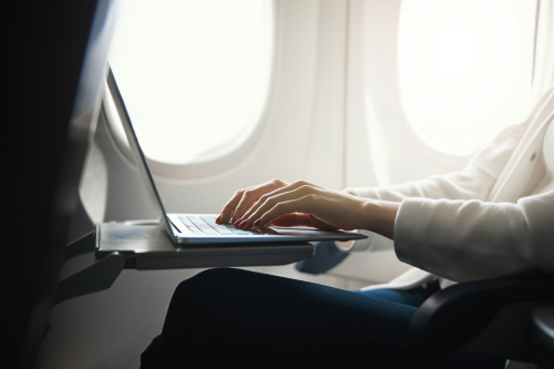 Person using a laptop while seated on an aeroplane, with hands typing on the keyboard and aircraft windows visible in the background