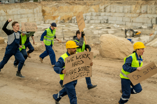 Construction workers in high-visibility clothing walk through a quarry-like worksite carrying protest signs during an industrial strike.