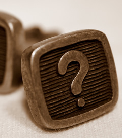 A close-up, sepia-toned image of a pair of vintage-style cuff links. The front of one cufflink is clearly visible, featuring a prominent question mark embossed in the center with a ridged background.