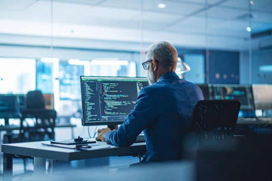 Photo of a senior software developer at work sitting at a desk wearing a blue shirt and glasses