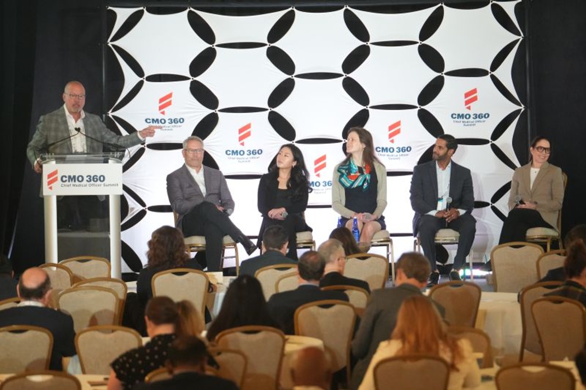 A panel discussion takes place at the CMO360 Summit in Boston, with industry leaders sharing insights on stage while delegates from across the biopharmaceutical sector watch from the audience. The event brings together executives to discuss clinical development, manufacturing, and commercial innovation.