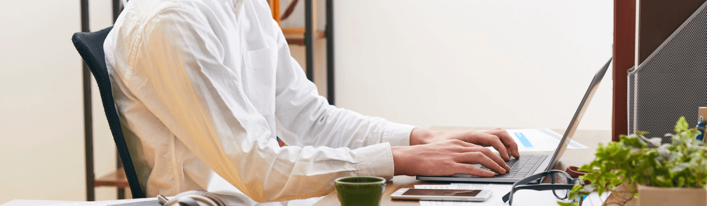 A man in a white shirt is typing on his laptop, concentrating on his work in a bright, organized workspace.