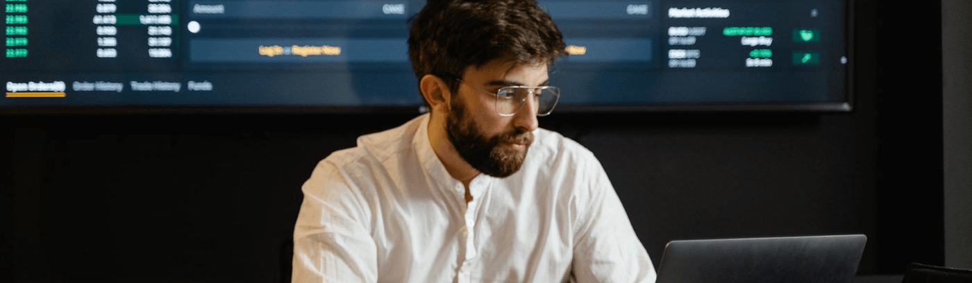 A bearded man wearing glasses is seated at a desk, focused on his laptop while working intently.