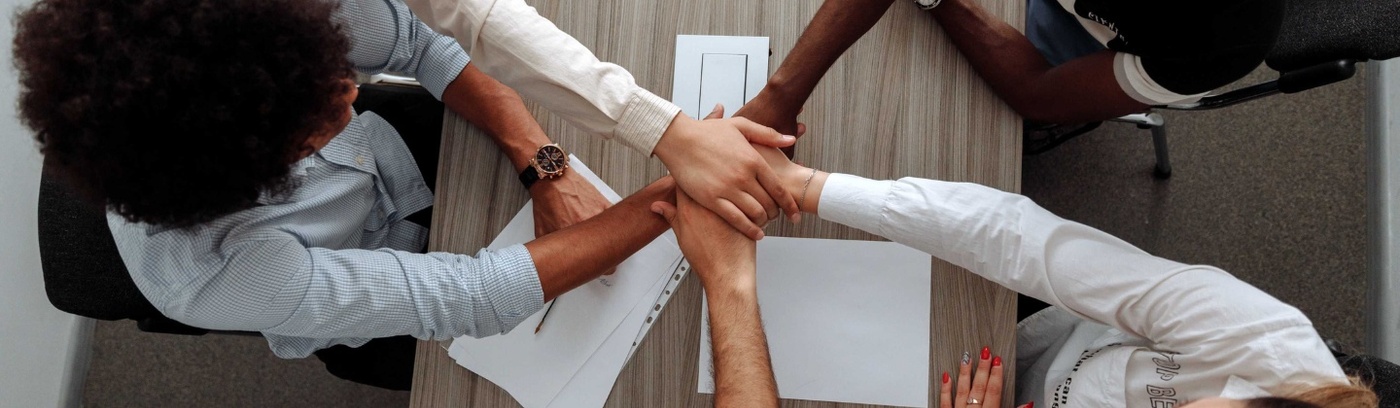 Four people at a table, hands raised and layered on top of each other, indicating enthusiasm or participation.